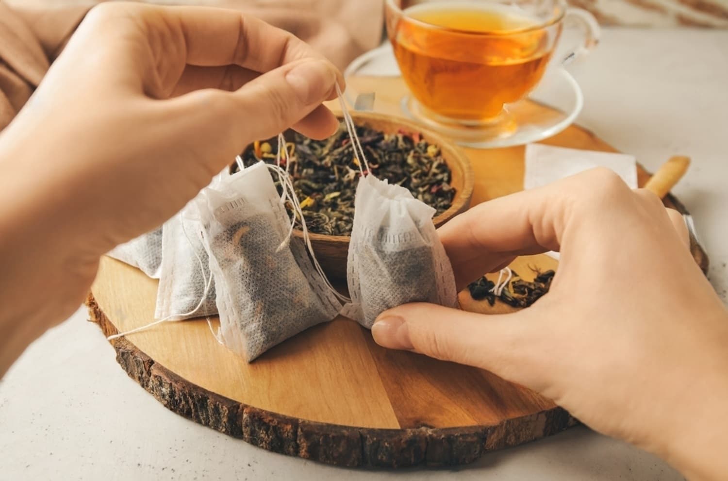 Hands preparing tea bags with loose leaf tea and a cup of brewed tea on a wooden board.