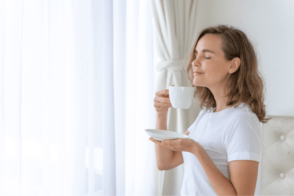 Woman enjoying tea by a window, eyes closed, wearing a white shirt.