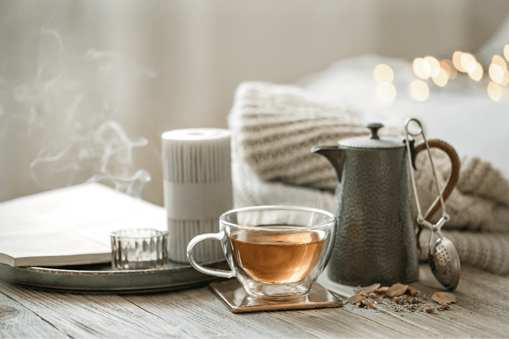 Steaming cup of tea with a teapot, candle, and open book on a cozy wooden table.