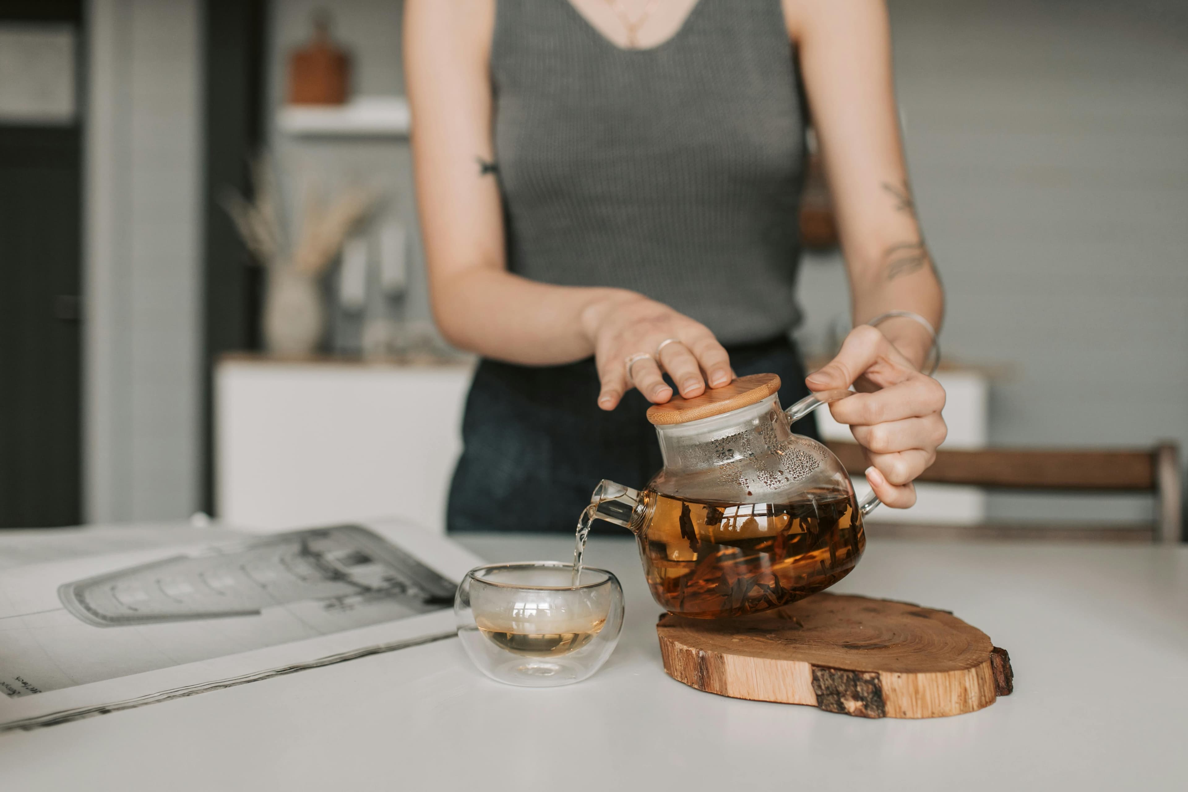 Person pouring herbal tea from a glass teapot into a cup on a wooden coaster.