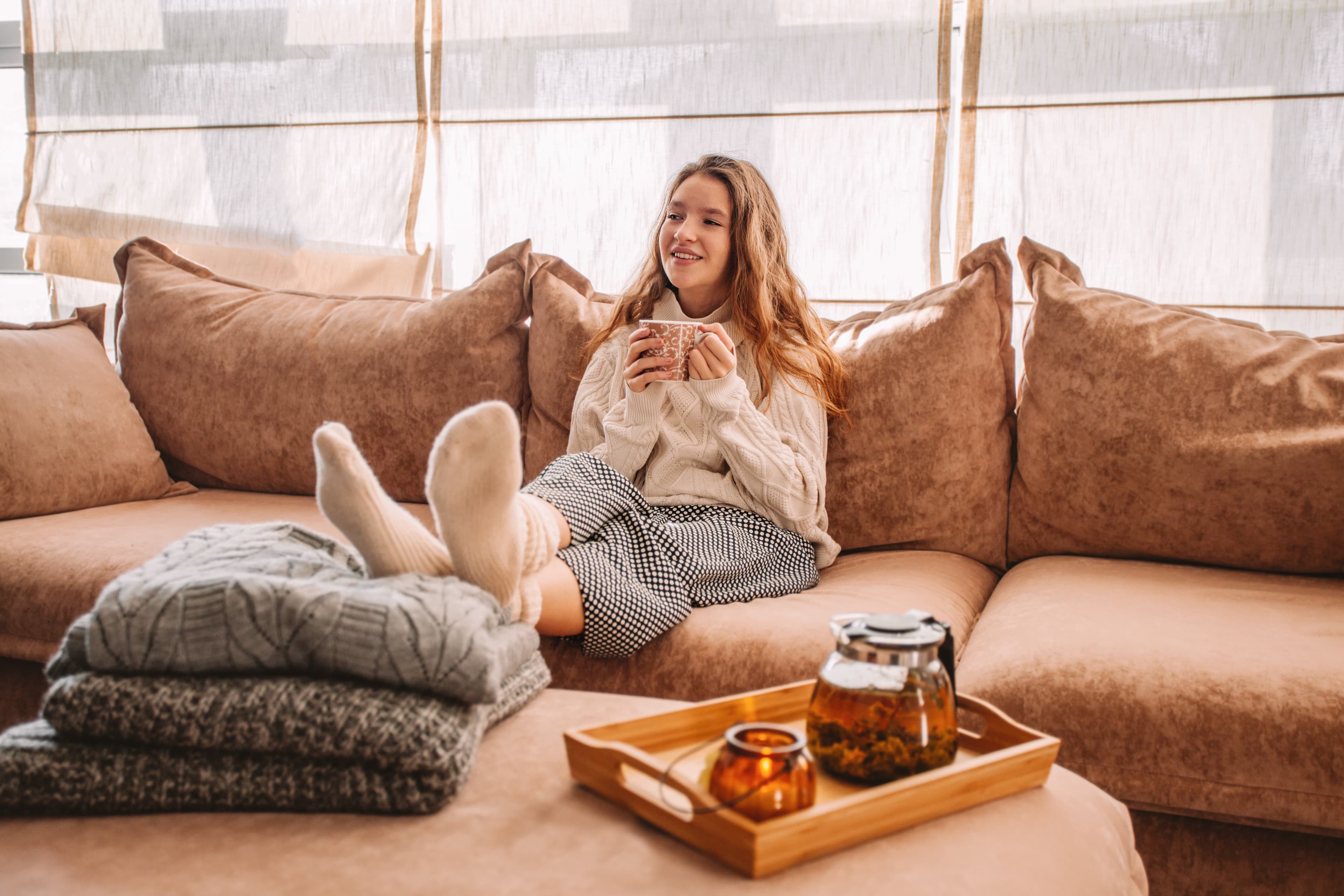 Woman relaxing on a sofa with a cup of tea, next to a tray with a teapot and candles.