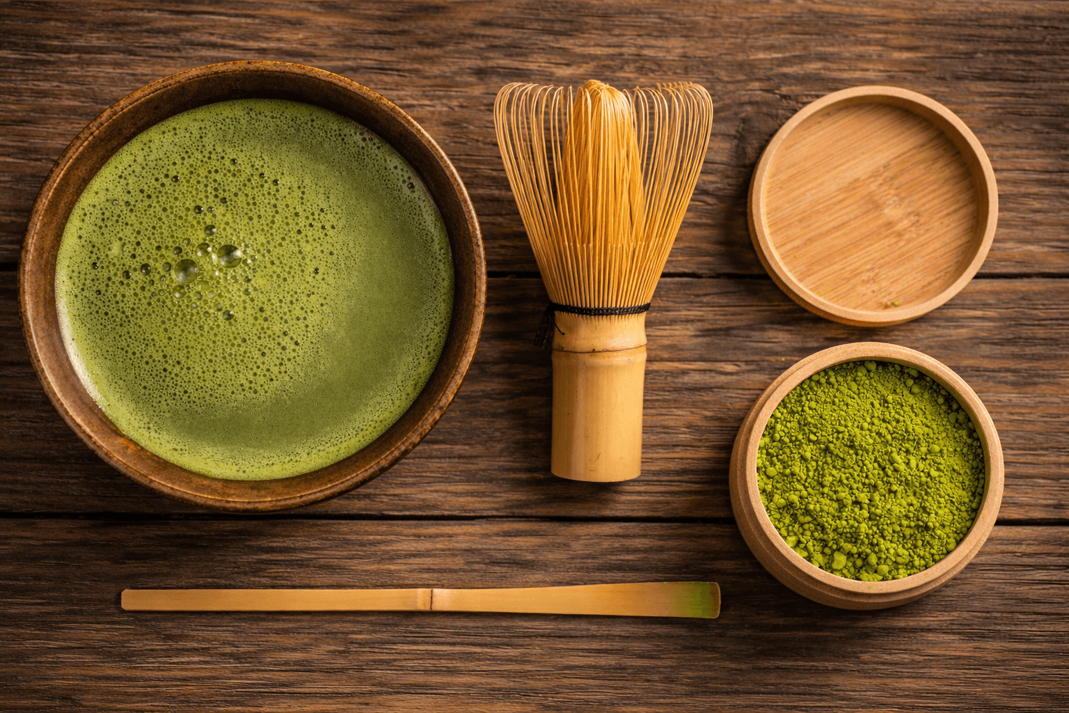 Matcha tea set with a bowl of frothy matcha, bamboo whisk, powder container, and scoop on wooden table.